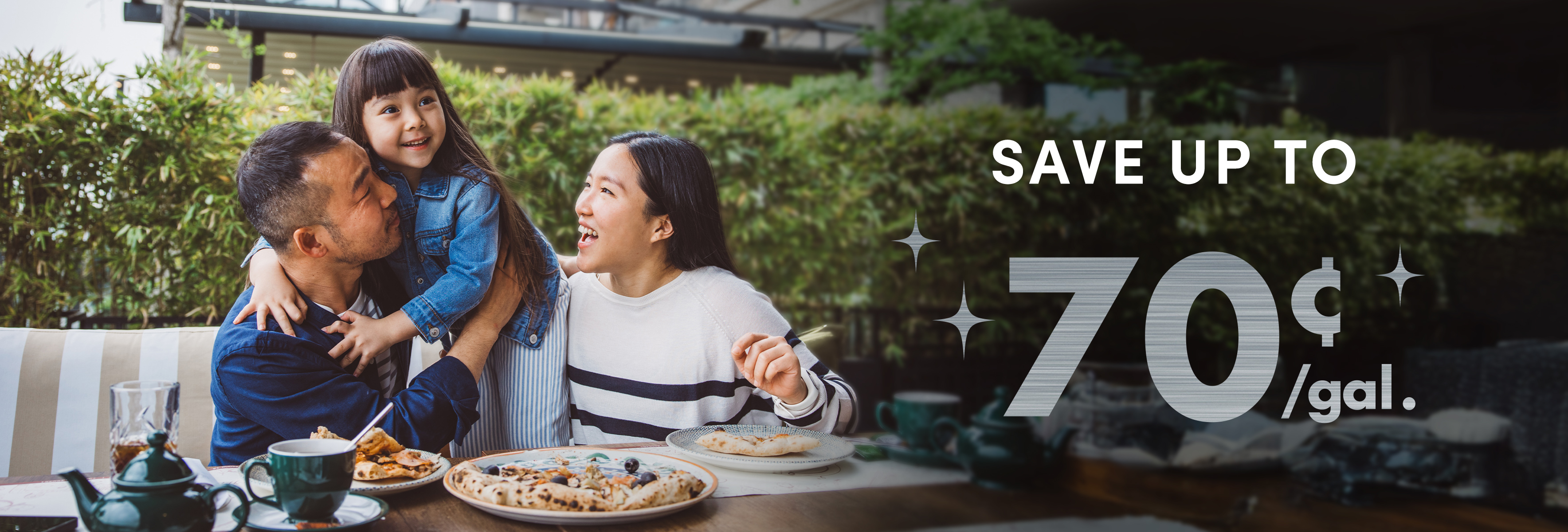 A family enjoys a meal outdoors, surrounded by greenery, with dishes of food and drinks on the table. A promotional text reads 'Save up to 70¢/gal.'